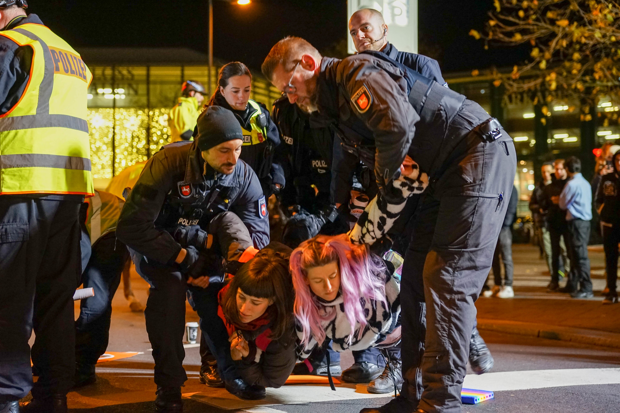 A photograph, taken at nighttime on a road with lampposts. It shows two young female climate activists being pulled off the ground by police in riot uniforms, surrounded by more police officers.