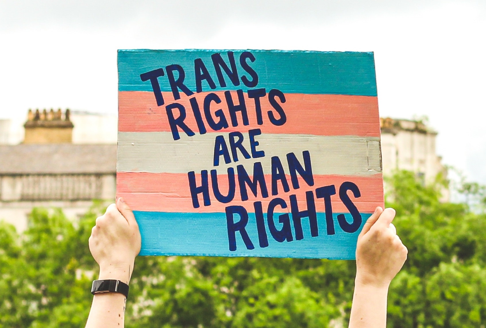 Een foto van een protestbord dat in de lucht wordt gehouden. Op het bord zijn de kleuren van de trans vlag geverfd met daarop de tekst Trans Rights are Human Rights.