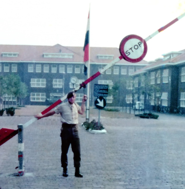 A person in uniform, holding up a boom barrier with a stop sign on it, with in the background a military base and a flag pole with the Dutch flag.
