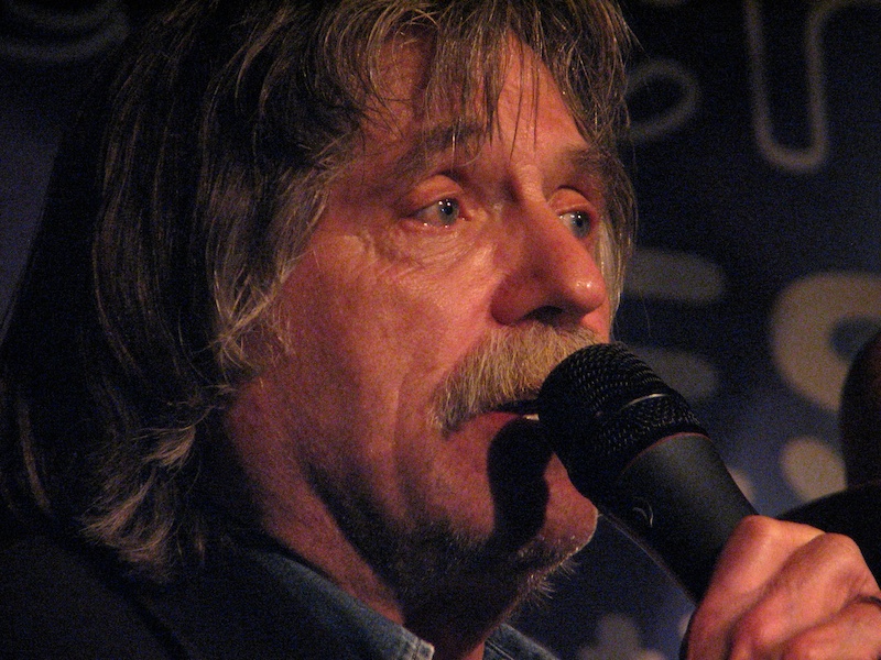 Een close-up foto van het gezicht van Johan Derksen. Hij staat in een donker verlichte ruimte, kijkt naar rechts en houdt een microfoon vast waar hij in spreekt.
