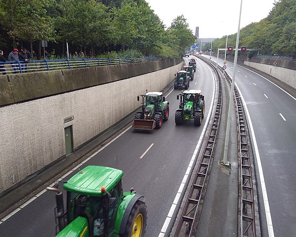 Een foto van een snelweg, genomen van boven. Zes groene tractors rijden over de snelweg in een lijn richting de camera. De andere weghelft is leeg, met in de verte borden dat de weg is gesloten.