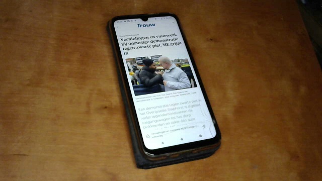 A photo of a smartphone laying on a wooden surface. On the screen is an article from newspaper Trouw, with a Dutch headline about Zwarte Piet followed by a photo of a black man and an older white man standing outside and talking.