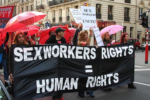 Five people walking at the front of a march. They are holding a large black banner which says â€œsex workers rights equals human rightsâ€� in bold white text. One person is holding a white sign that says â€œSex workers unite!â€�. People behind them are holding red umbrellas.