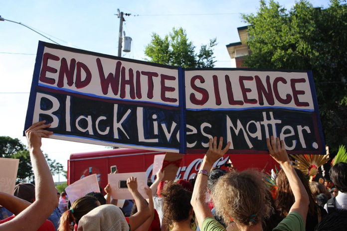 A cardboard sign being held up at a protest, reading 'End white silence. Black lives matter.'.