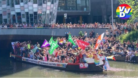 Een foto van een pride boot. De boot vaart door een kanaal. Er hangt een spandoek aan de zijkant. De boot staat helemaal vol met mensen, en er wordt gezwaaid met vlaggen van de PvdA, D66, Volt en de VVD. In de achtergrond is een kade te zien, ook helemaal vol met mensen.