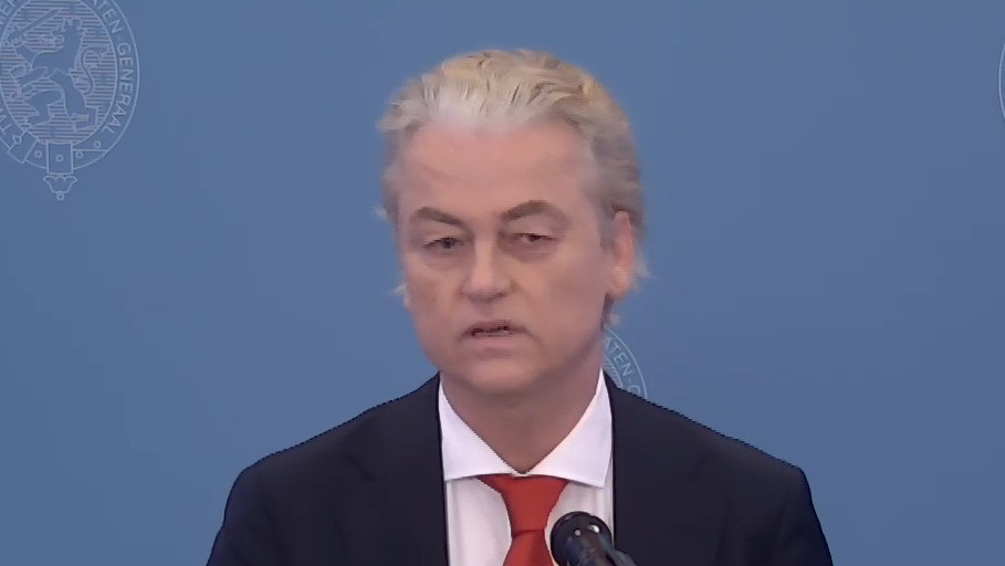 Een foto van Geert Wilders, staande bij een glazen spreekgestalte voor een blauwe achtergrond, tewijl hij het hoofdlijnenakkoord presenteert.